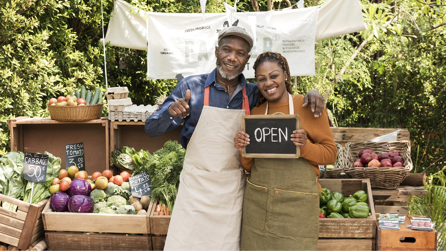A happy couple in aprons stands in front of a produce stand. He has his arm over her shoulder and is giving a thumbs up while she holds a small chalkboard reading 'Open.'