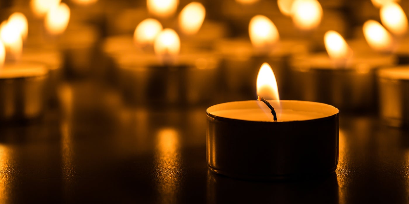 At a Catholic Charities housing site, a man with a beard in a fleece vest bends over a washing machine, checking on his clothes. Another man, wearing a purple hoodie, sits nearby.