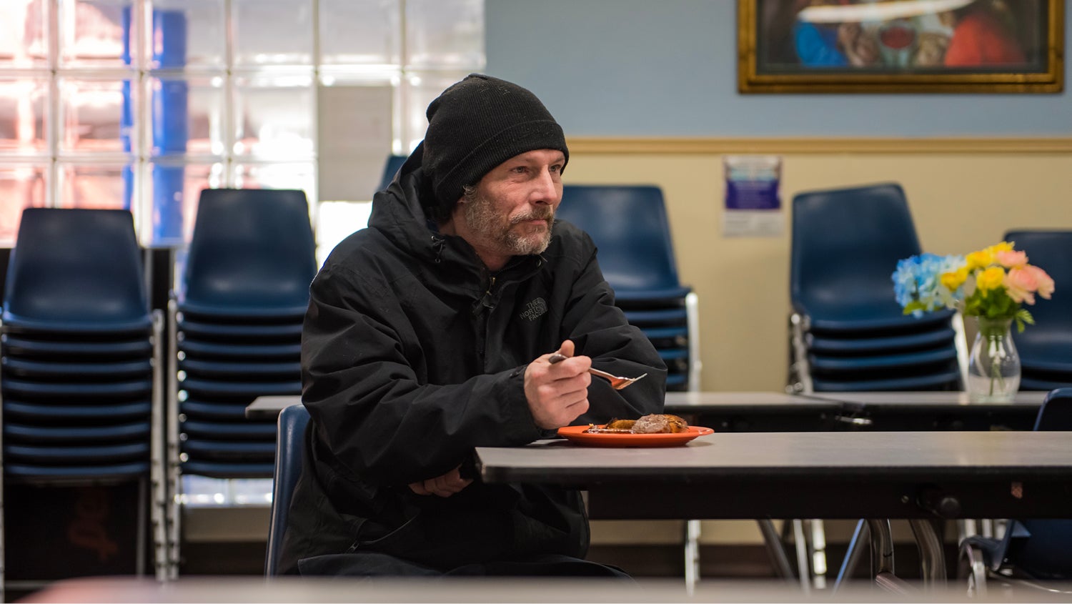 A man in a dark hoodie and knit cap sits at a dining table with a plate of food at a Catholic Charities congregate dining center.
